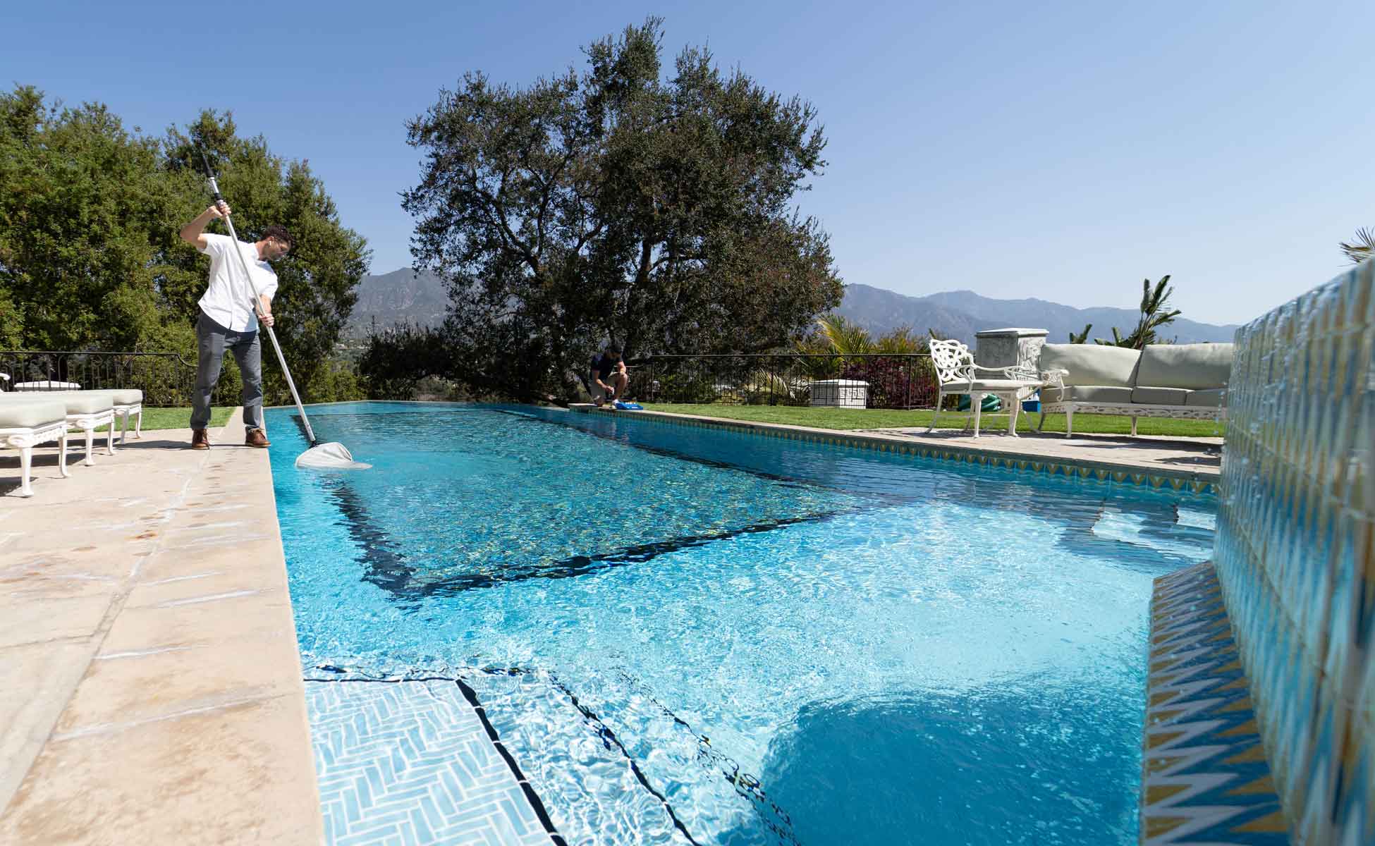 Pool Care LA technician netting the surface of a tiled hillside pool with the San Gabriel Mountains behind it