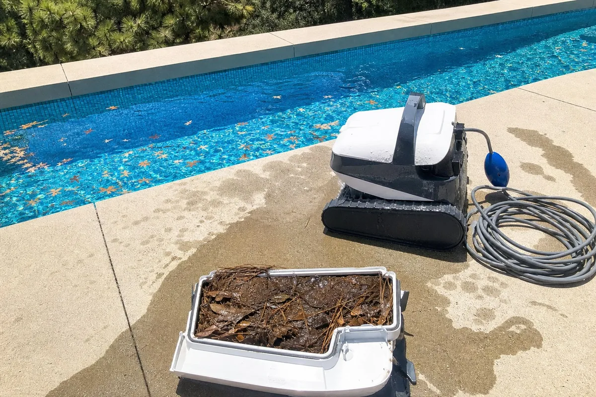 A robotic pool cleaner on the deck with its debris container pulled out and packed full of wet leaves and pine needles